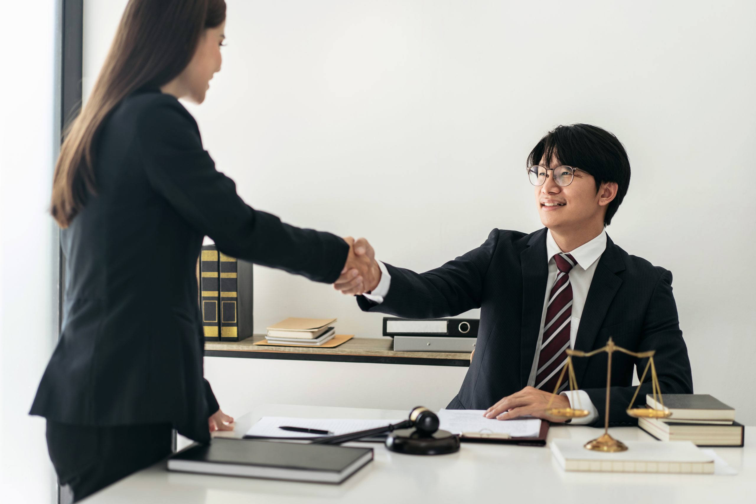 A man and woman in formal attire shake hands in an office. The man is seated at a desk with legal books, a gavel, and scales of justice, suggesting a professional legal setting. The scene hints at the conclusion of an expungement hearing, signaling hope for fresh beginnings.