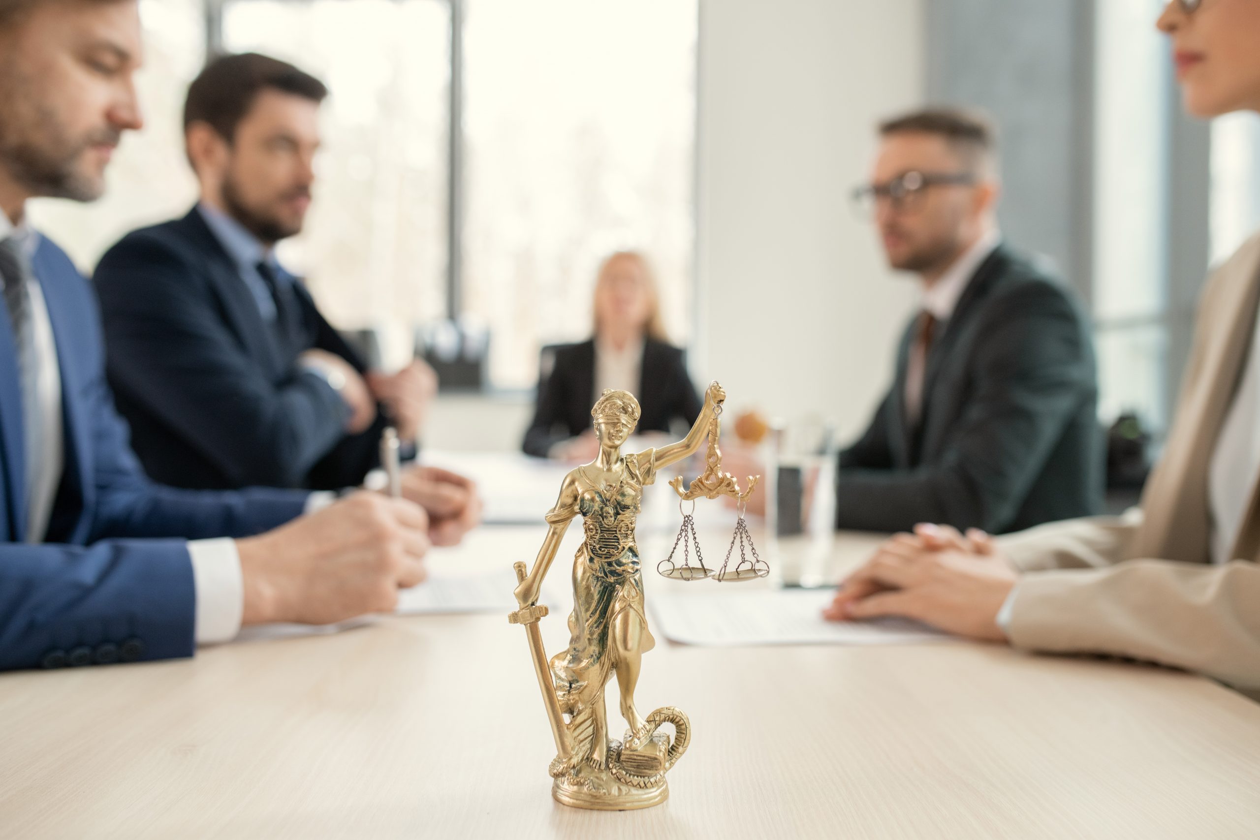 Focus on golden Themis with scales of justice and sward placed on lawyers table, colleagues sitting across from each other in background