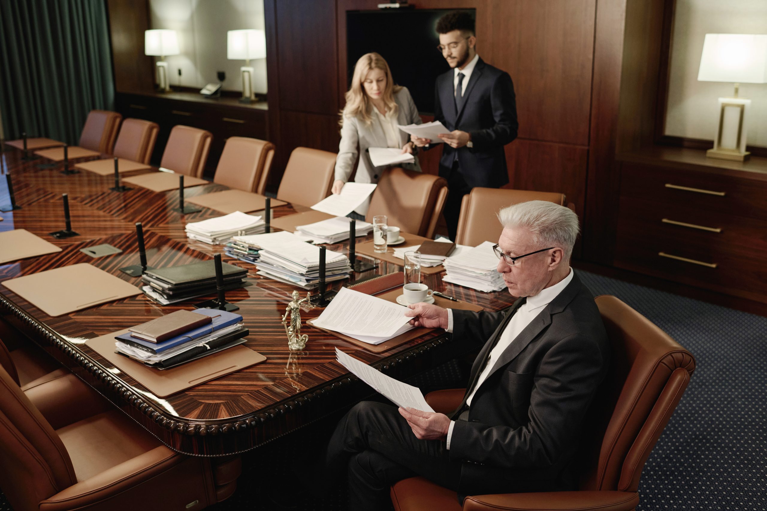 A man in a suit sits at a large conference table, reviewing personal injury documents. A young man and woman stand nearby, going over insurance papers. The table is cluttered with stacks of documents, folders, and coffee cups. The room is warmly lit and has a formal atmosphere.