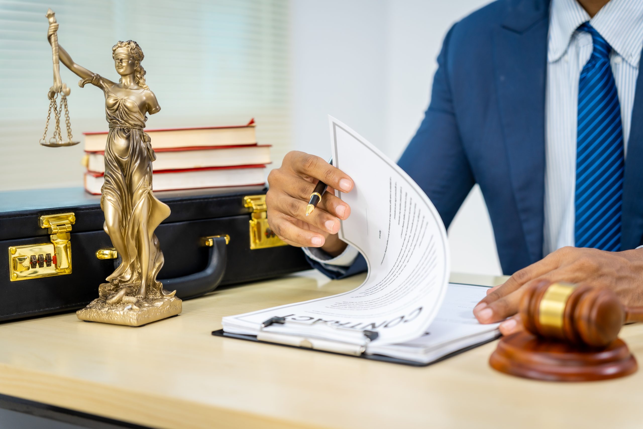 A person in a suit efficiently reviews a contract on a desk adorned with a gavel, Lady Justice statue, and stack of books. A black briefcase is also present. The setting suggests an office where best practices in the legal field are paramount.