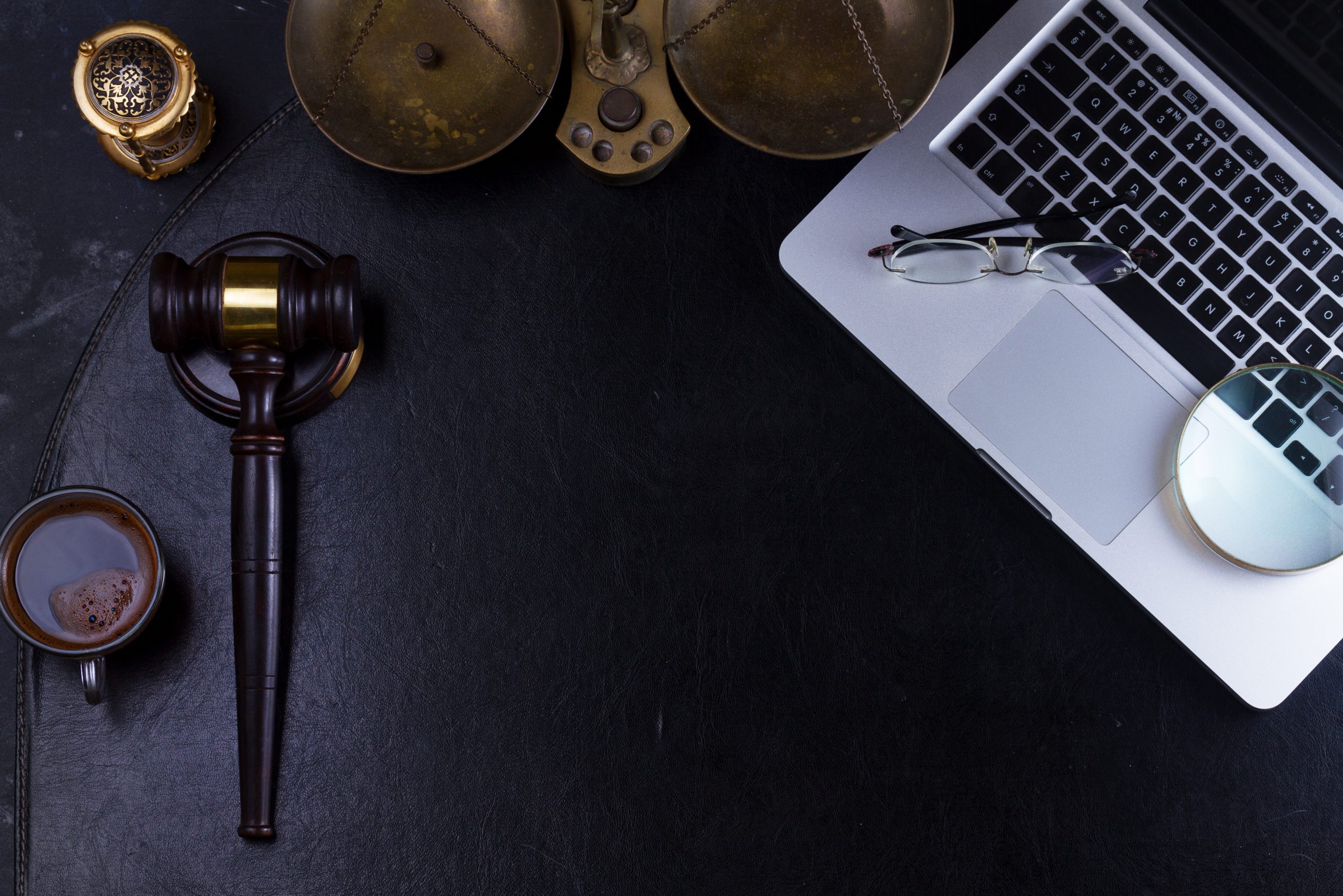 A laptop and reading glasses rest on a dark desk, accompanied by lawsuit statistics, a magnifying glass, a cup of coffee, a gavel, and a vintage scale.