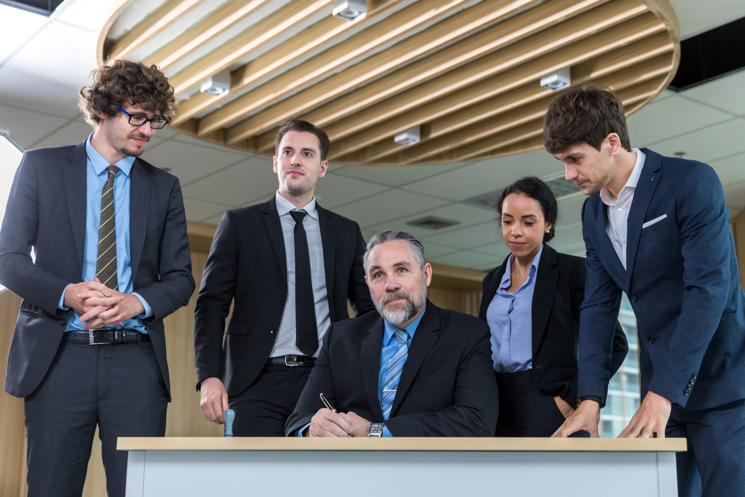 A group of five business professionals gathers in a conference room. A trial attorney seated in a suit signs a document at the table, as the others stand around, observing intently. The room boasts a modern ceiling design and large windows that cast natural light over their deliberations.