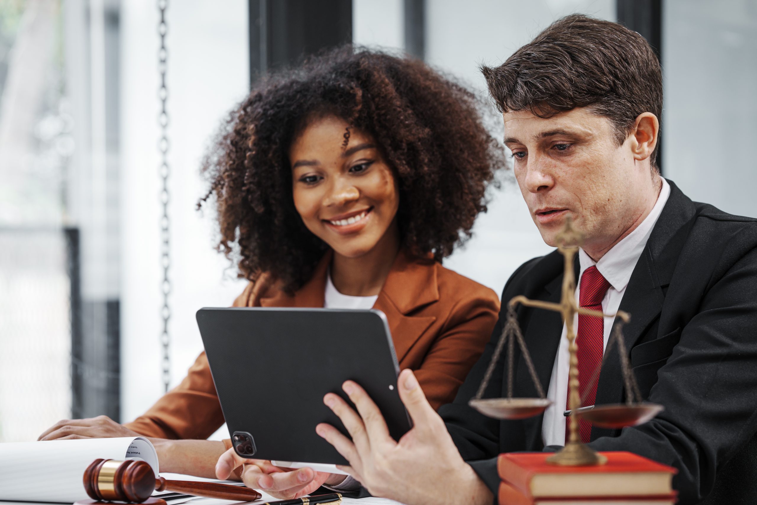 A woman and a man in business attire are sitting at a desk, looking at a tablet. The man is holding the tablet, and the woman is smiling, discussing the benefits of expungement. A gavel and balance scales accompany books and documents on the desk, symbolizing their path to a fresh start.